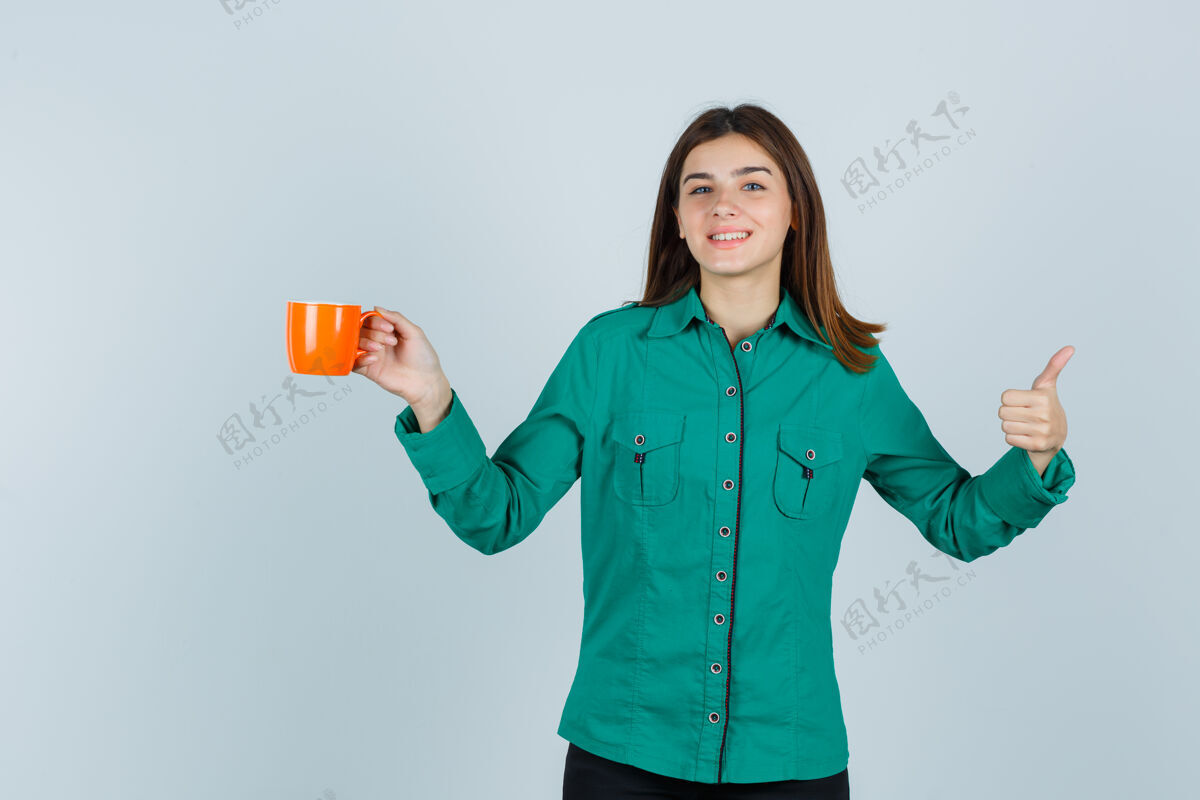 成人穿着衬衫的年轻女士手里拿着一杯橘色的茶 竖起大拇指 看上去很高兴 正前方的景色女孩人牙齿
