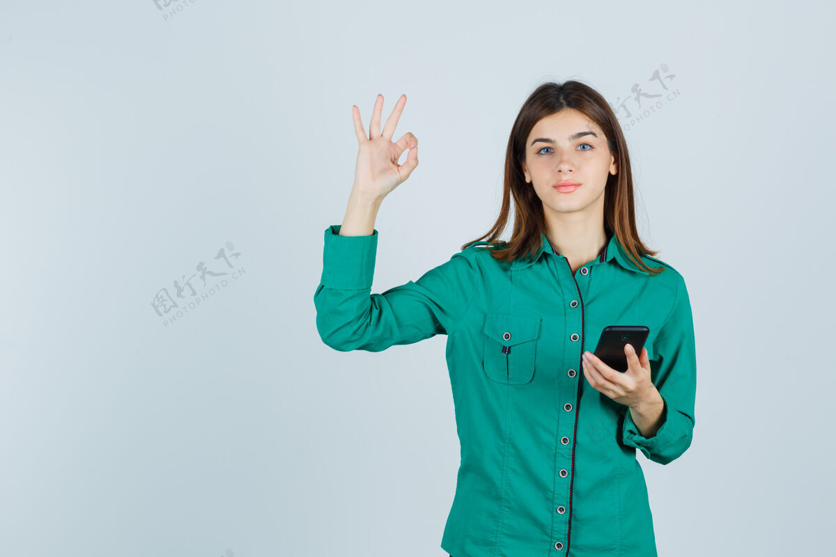 手机手持手机的年轻女士的肖像 在绿色衬衫中显示ok手势 看起来很高兴的正面视图黑发女孩女性