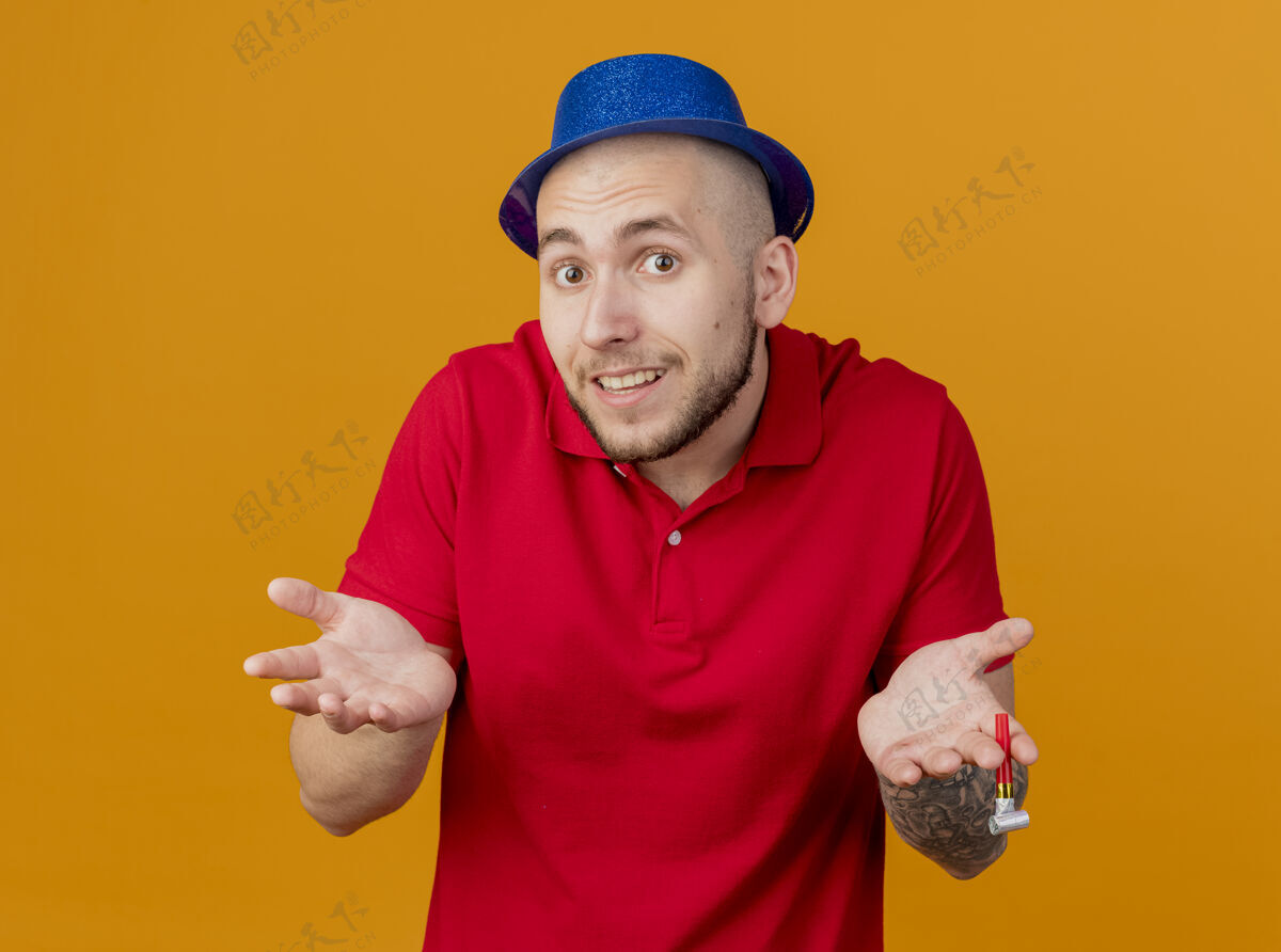 男人无知的年轻英俊的斯拉夫党人戴着党的帽子看着相机显示空手孤立在橙色背景表演无知手