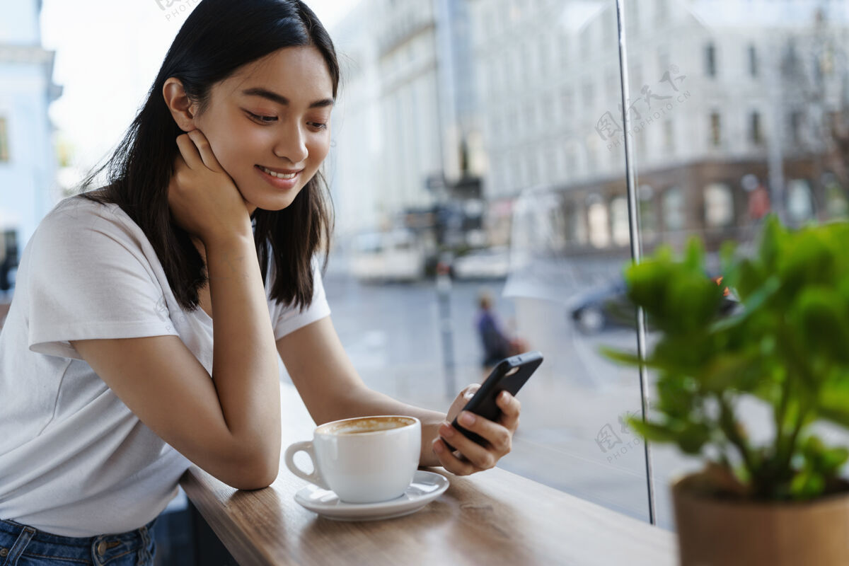 杯子在靠近窗户的餐厅里 一个女孩一边用智能手机聊天 一边喝咖啡 一边对着手机屏幕微笑城市年轻人桌子