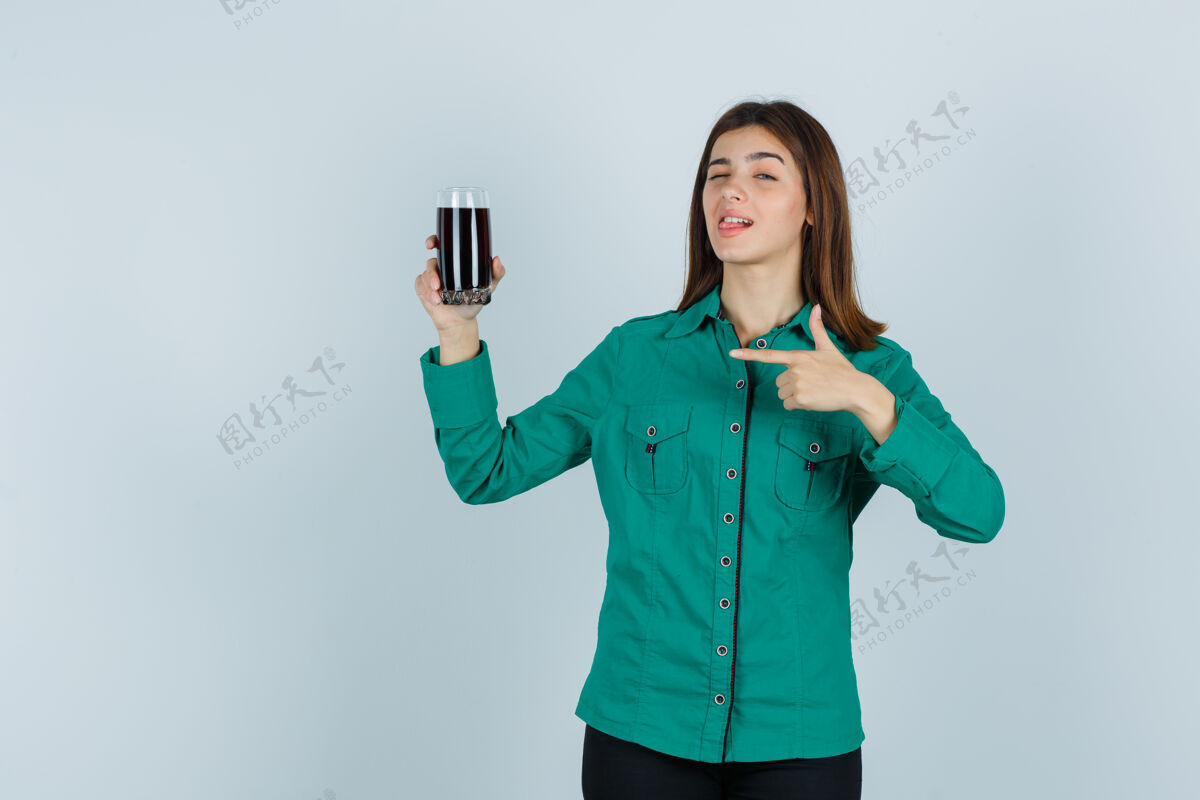 肖像年轻女孩穿着绿色上衣 黑色裤子 手里拿着一杯黑色液体 指着它 眨着眼睛 看起来很可爱 前视图衬衫指着室内