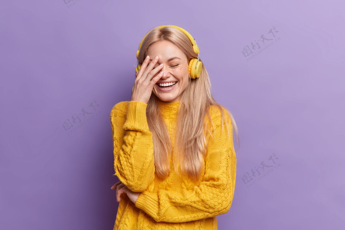 紫色喜出望外的欧洲年轻金发女子大声笑出声来让掌心通过无线耳机听音乐穿着休闲的黄色毛衣套头衫毛衣穿着