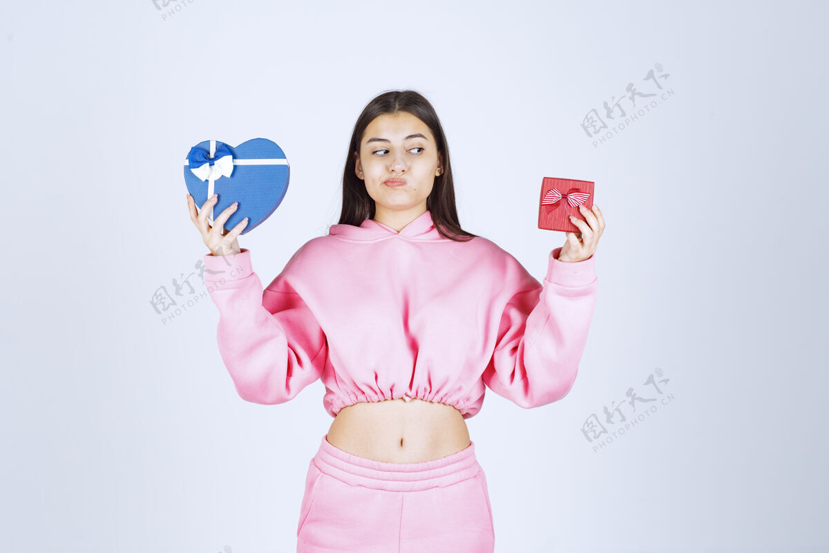 怀疑穿着粉色睡衣的女孩拿着红色和蓝色心形的礼品盒 在它们之间做出选择工人促销模特