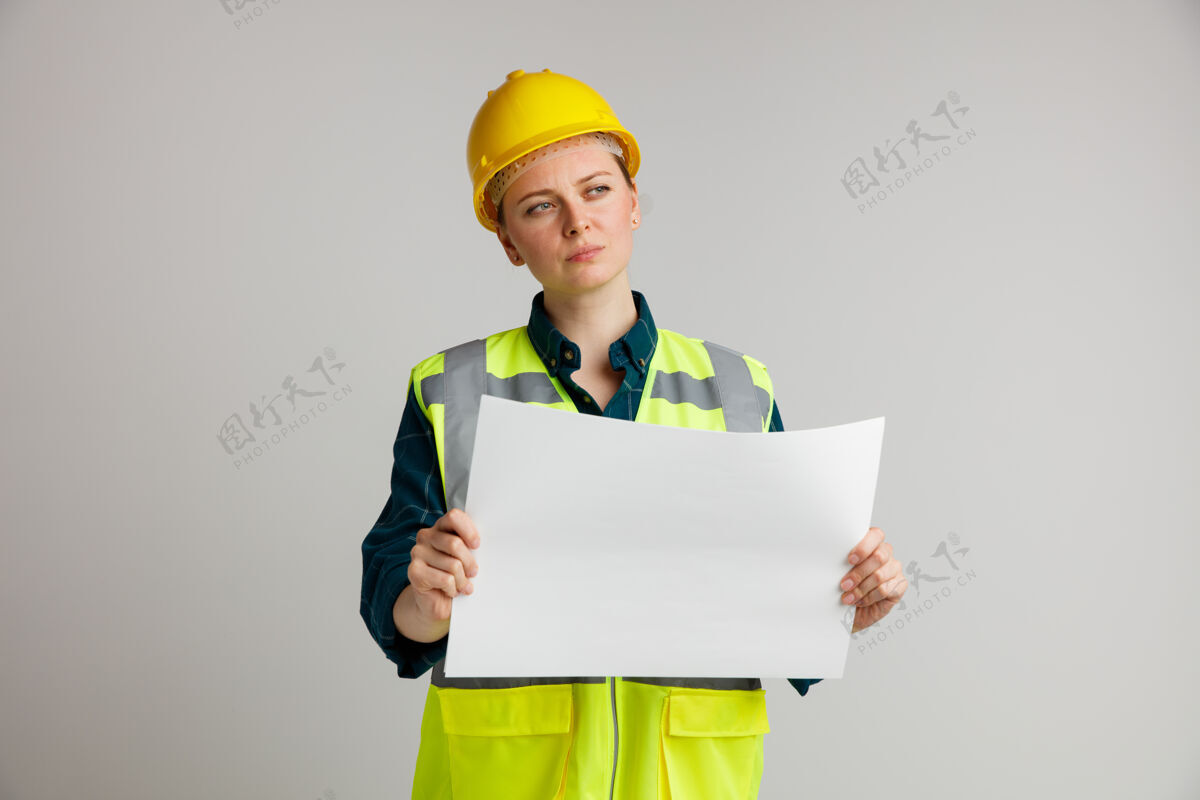 穿困惑的年轻女建筑工人戴着安全帽和安全背心拿着纸看着身边建筑纸女
