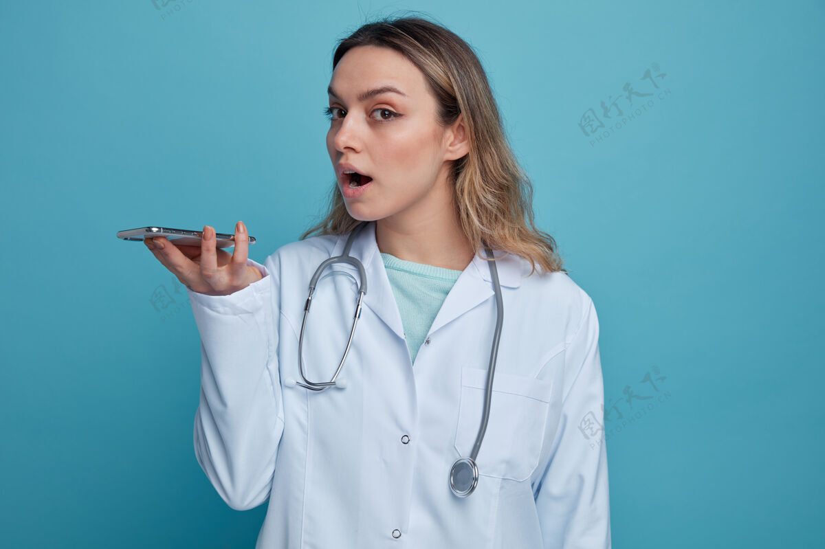 医疗令人印象深刻的年轻女医生穿着医用长袍 脖子上戴着听诊器 手持手机 通过麦克风水平通话手机印象脖子
