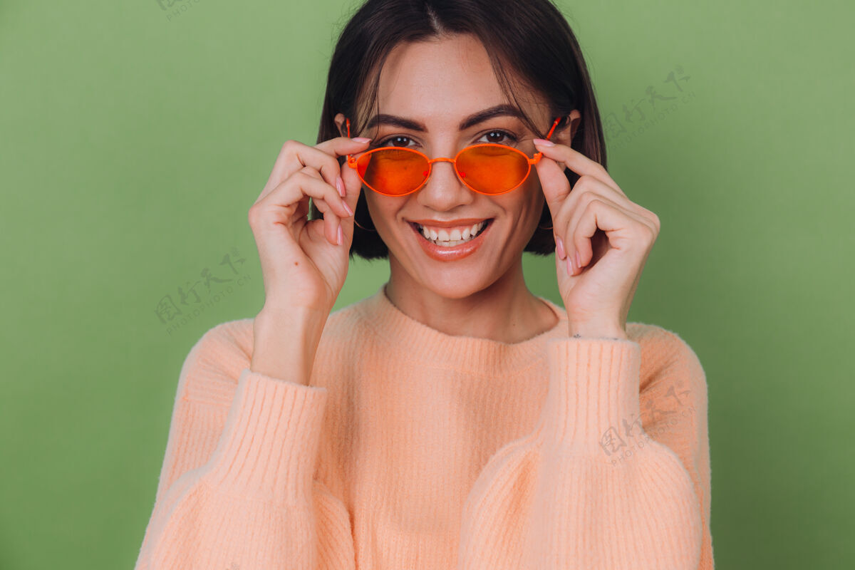 快乐年轻时尚的女士穿着休闲的桃色毛衣 戴着橙色的眼镜 隔着绿橄榄色的墙壁 开心的 积极的笑着 围着复制空间转眼镜女孩可爱