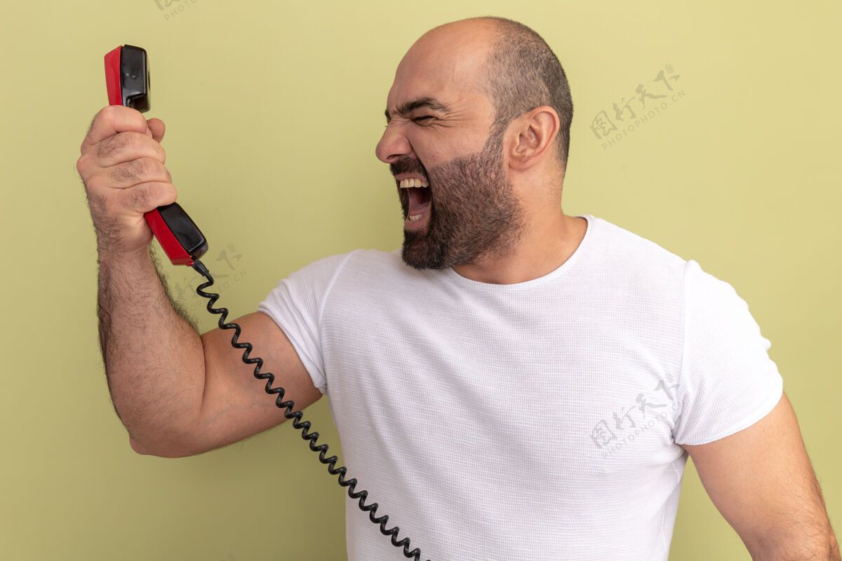 站一个留着胡子 穿着白t恤的男人拿着旧电话站在绿色的墙上大喊大叫电话好斗男人