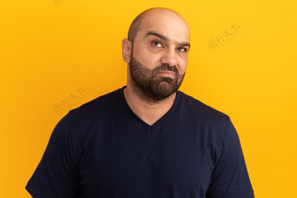 站着一个不高兴的留着胡子的男人 穿着海军蓝t恤 站在橘色的墙上 抬起头苦笑着 带着失望的表情不高兴失望做着