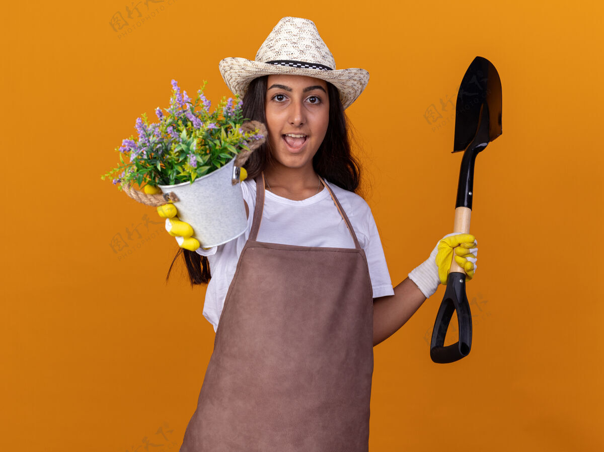 年轻年轻的园丁女孩拿着盆栽和铲子站在橙色的墙上开心地笑着欢呼花园站