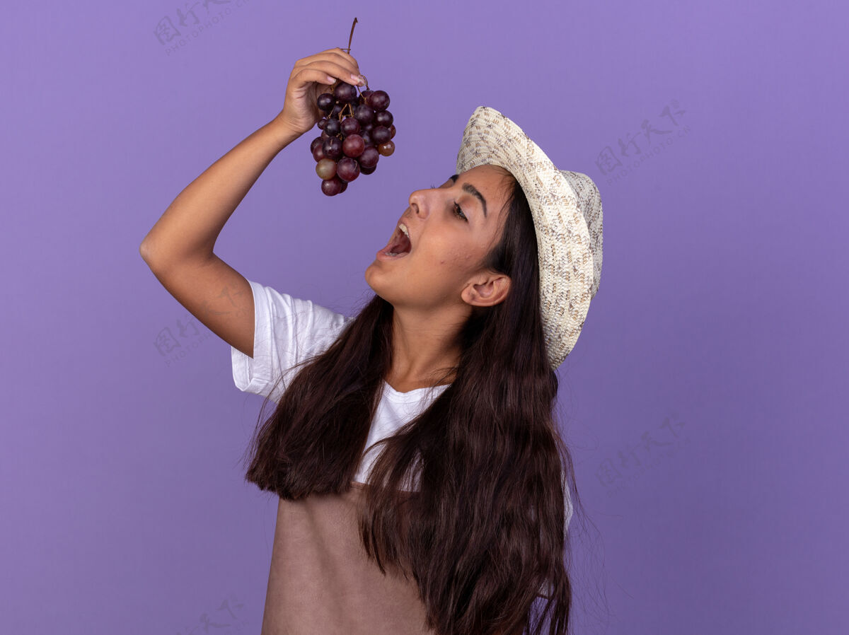 味道穿着围裙 戴着夏帽 嘴里叼着一束葡萄的年轻园丁姑娘站在紫色的墙上要尝一尝立场围裙帽子