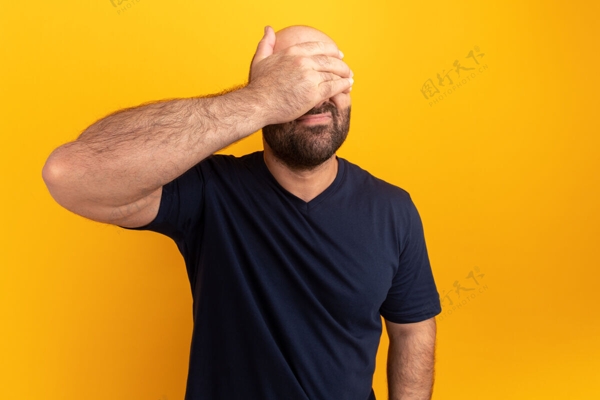掩护留着胡子的男人穿着海军蓝t恤 看起来很疲惫 无聊 用手遮住眼睛站在橙色的墙上表情男人胡子