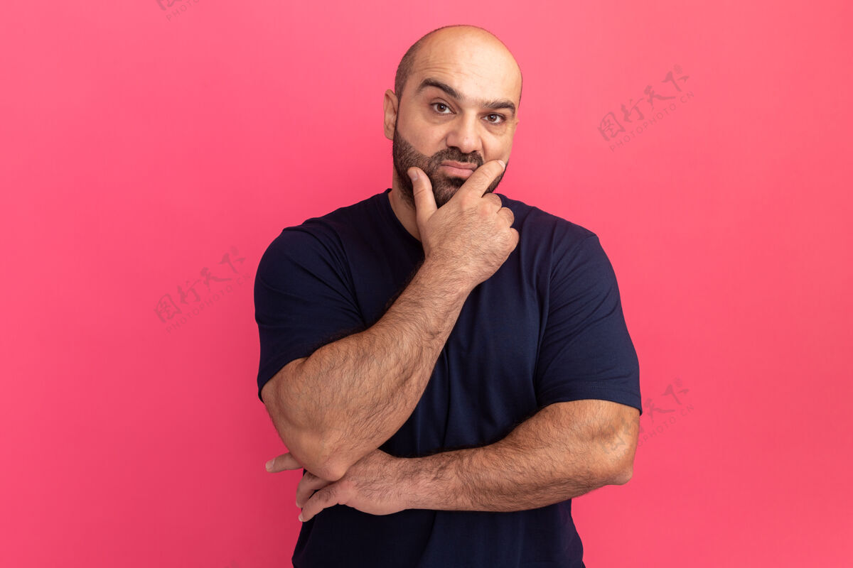 站立留着胡子的男人穿着海军t恤 手放在下巴上 站在粉色的墙上思考思考胡须下巴