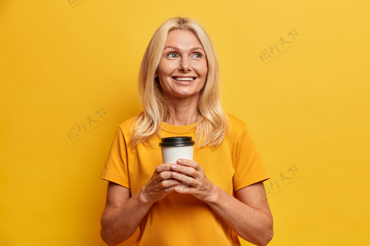 老快乐的满脸皱纹的金发女人化妆有度假计划喜欢喝外卖咖啡穿着休闲的黄色t恤摆出室内思考家庭的姿势人们的年龄和业余时间的概念想象皱纹放松