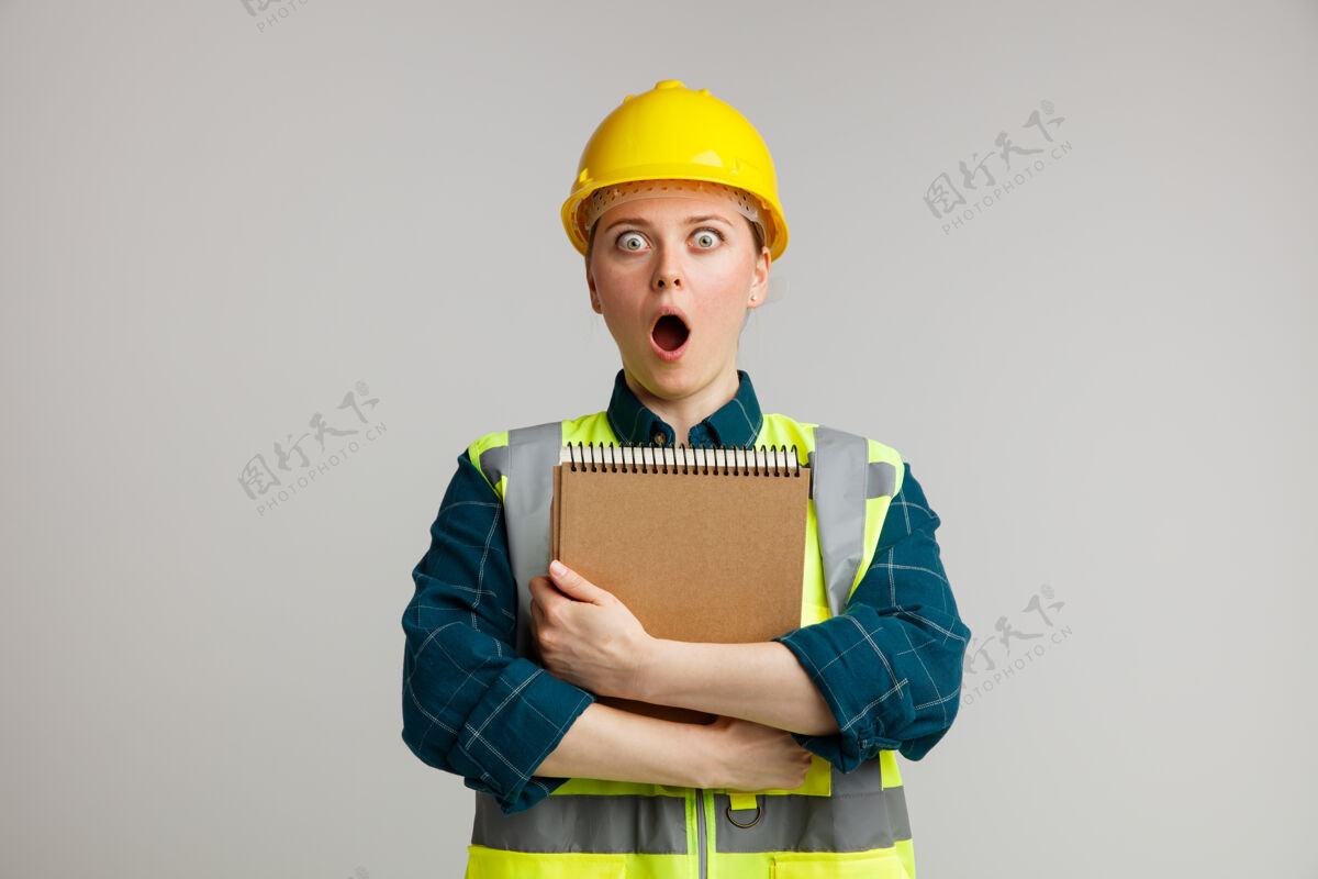 拥抱惊讶的年轻女建筑工人戴着安全帽和安全背心拥抱记事本惊喜建筑记事本