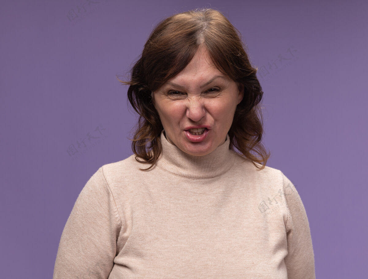 愤怒穿着米色高领毛衣的中年妇女站在紫色的墙上怒气冲冲站着中年年龄