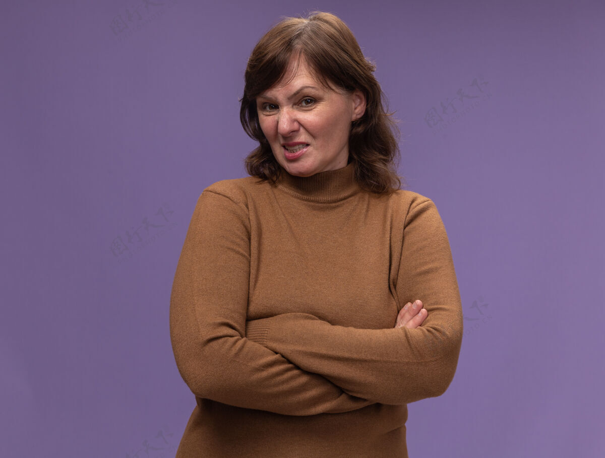 恼怒穿棕色高领毛衣的中年妇女站在紫色的墙上 双臂交叉 既恼火又恼火高领毛衣恼怒交叉