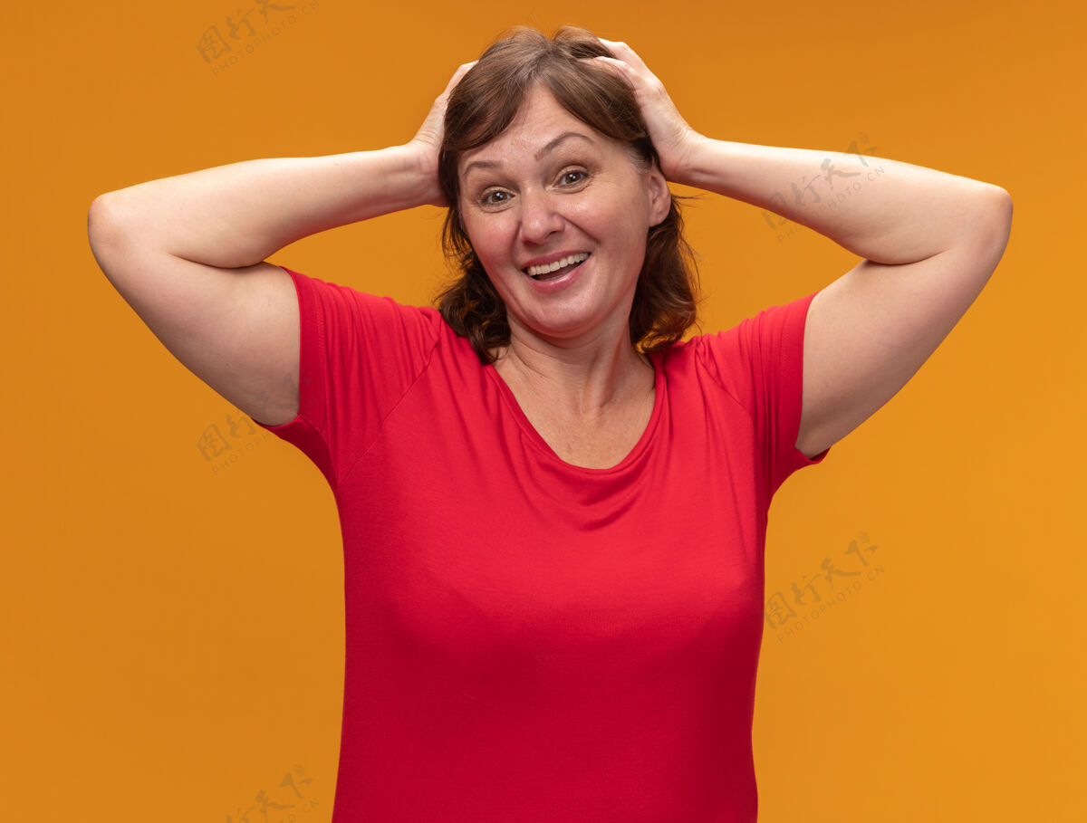 积极身穿红色t恤的中年妇女 双手放在头上 站在橙色的墙上 快乐而积极头立场年龄