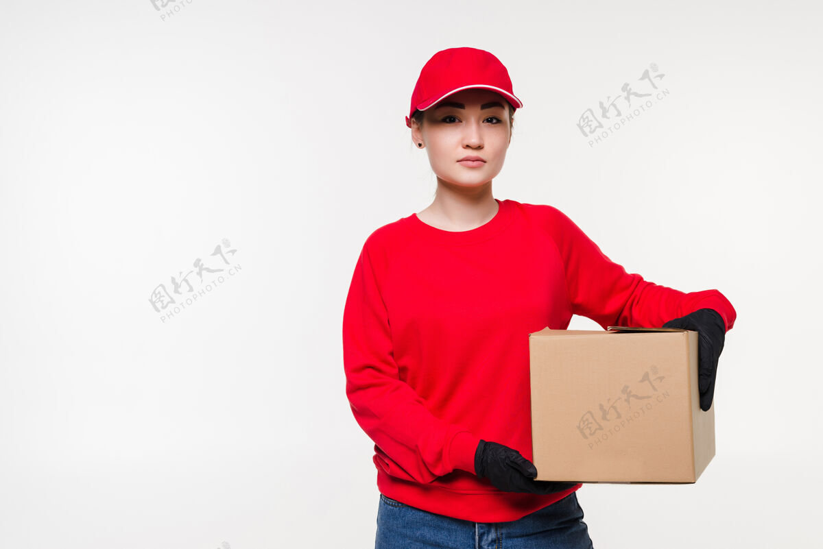 面罩一名身穿红色制服的女送货员被隔离在白色的墙上快递员戴着医用手套 戴着帽子 穿着红色t恤 作为经销商手持纸板箱送货收包裹检疫产品邮件