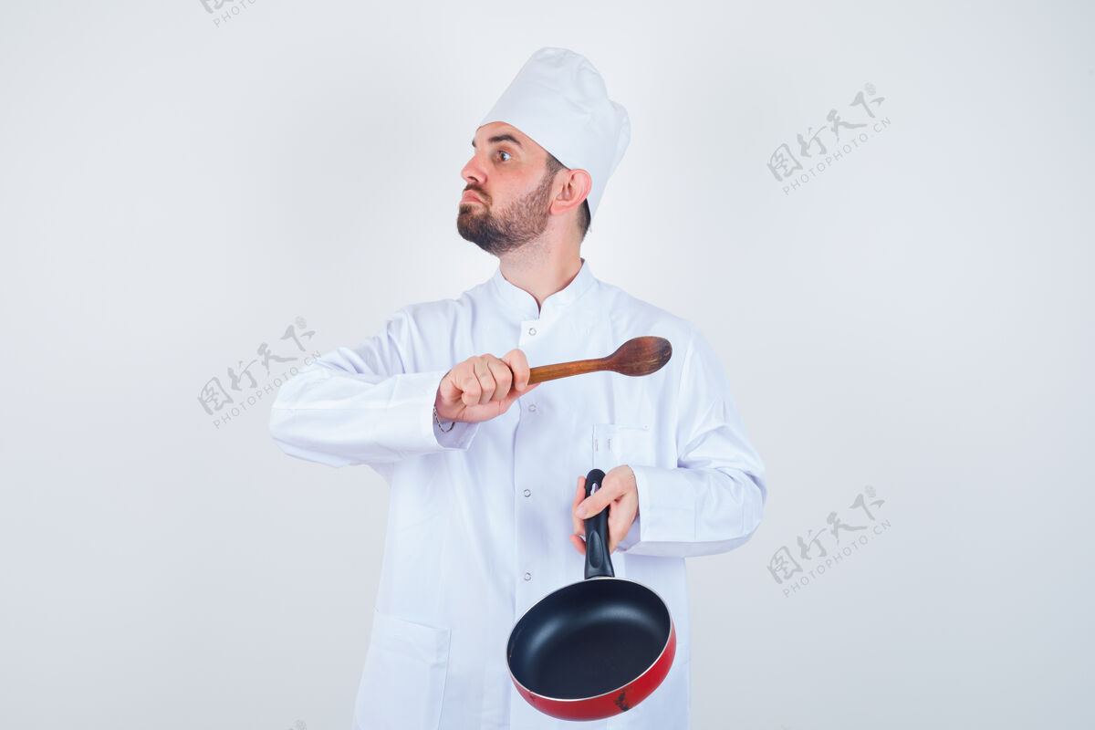制服一张年轻男厨师的画像 穿着白色制服 拿着煎锅和木勺威胁 看上去很紧张立场烹饪前面