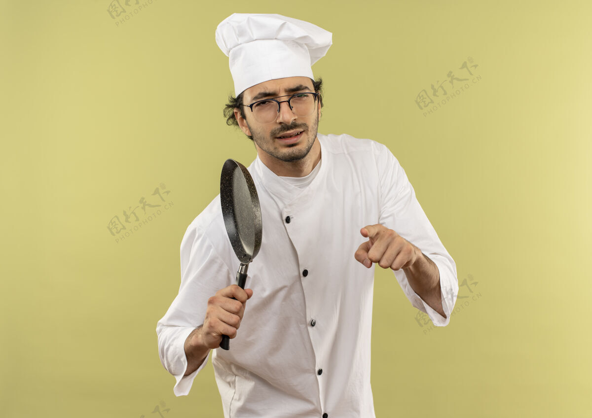 男性自信的年轻男厨师穿着厨师制服 戴着眼镜 拿着煎锅 向你展示手势显示平底锅年轻