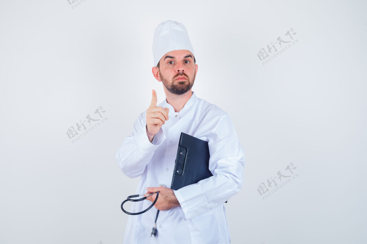 治疗年轻男医生拿着剪贴板 听诊器 穿着白色制服向上指着 看起来很聪明的前视图男性医院医生