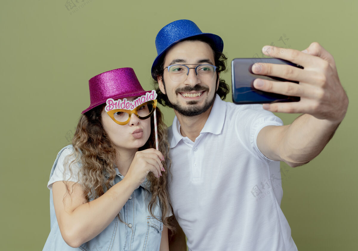 绿色高兴的年轻夫妇戴着粉红色和蓝色的帽子自拍和女孩举行化妆眼罩坚持帽子请手杖