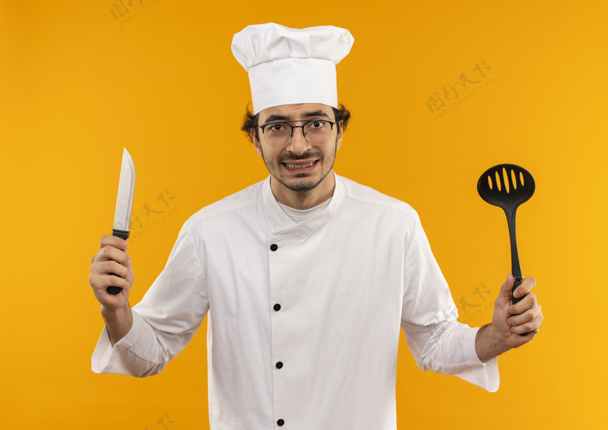 年轻年轻的男厨师穿着厨师制服 戴着眼镜 手里拿着抹刀和刀黄色眼镜刀