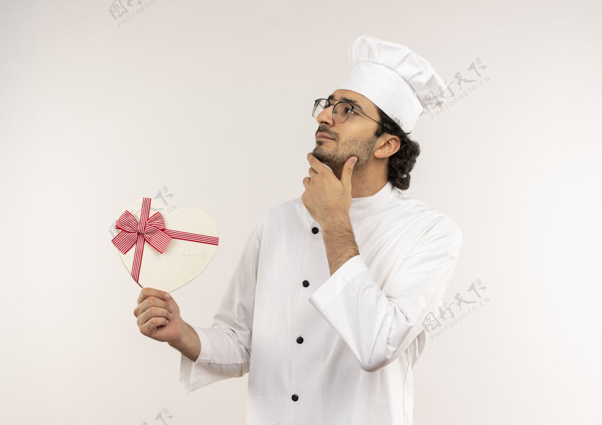 心看着一边想着的年轻男厨师 穿着厨师制服 戴着眼镜 手里拿着心形礼盒 手放在下巴上穿年轻厨师