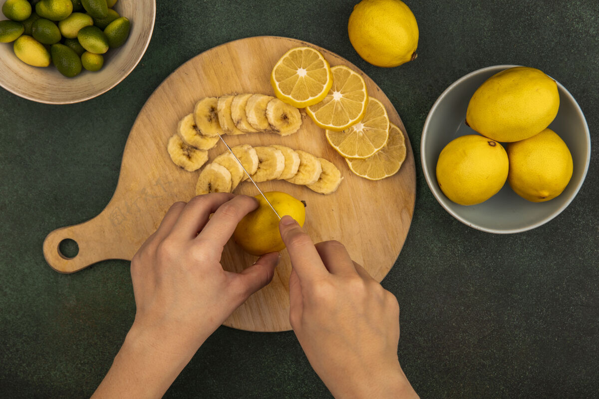 美味俯视图中的女性手切割新鲜的柠檬在一个木制的厨房板上用刀与金盏花在一个绿色的表面碗柠檬板女性