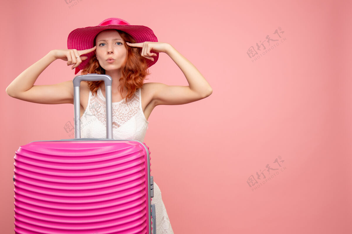 模特年轻的女性游客与粉红色的帽子和粉红色的墙壁上的包的正面视图肖像人度假