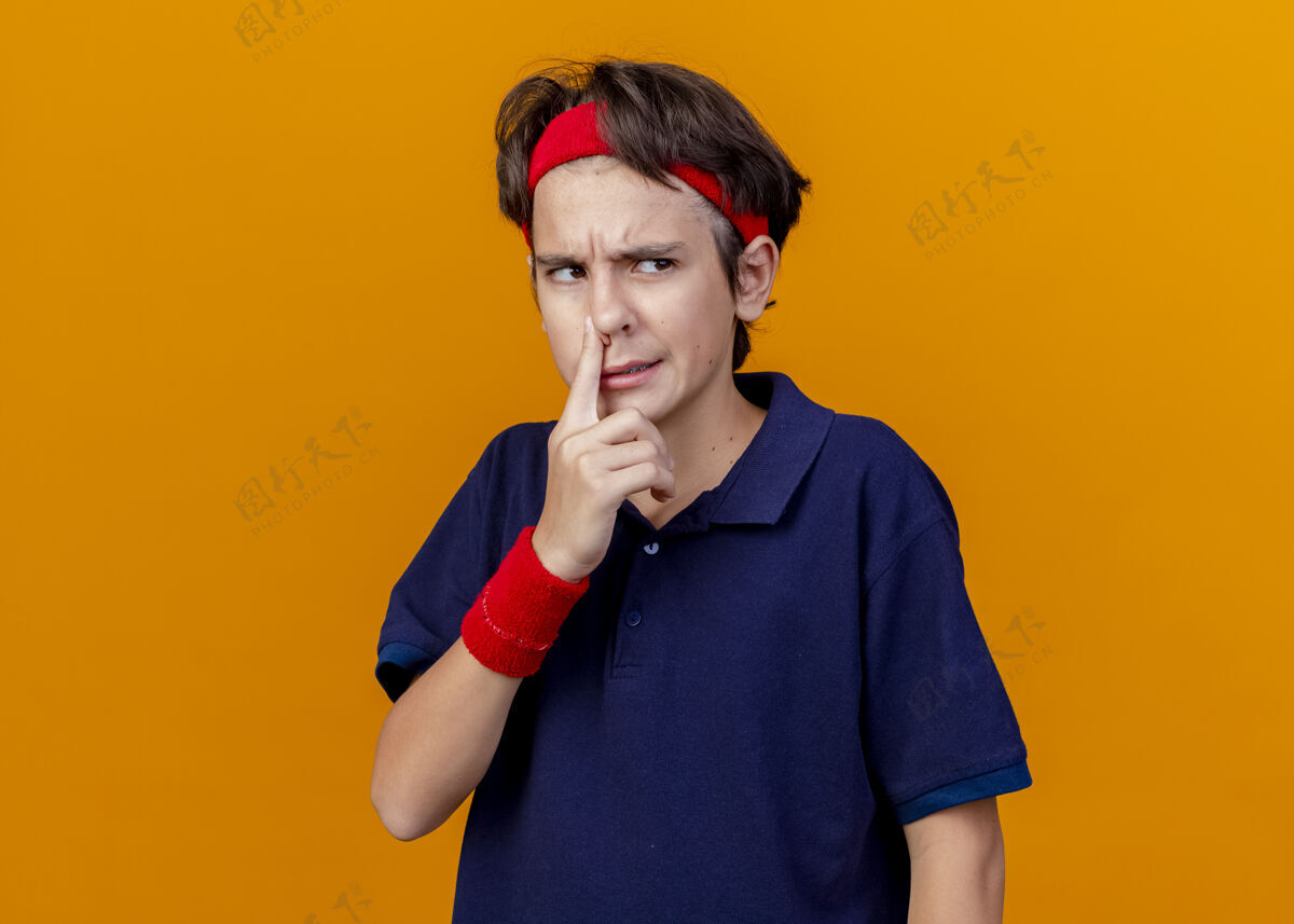 帅气恼怒的年轻英俊的运动男孩戴着头带和护腕 戴着牙套 看着一边把手指放在鼻子上 隔离在橙色的墙上 留着复制空间鼻子壁板手指