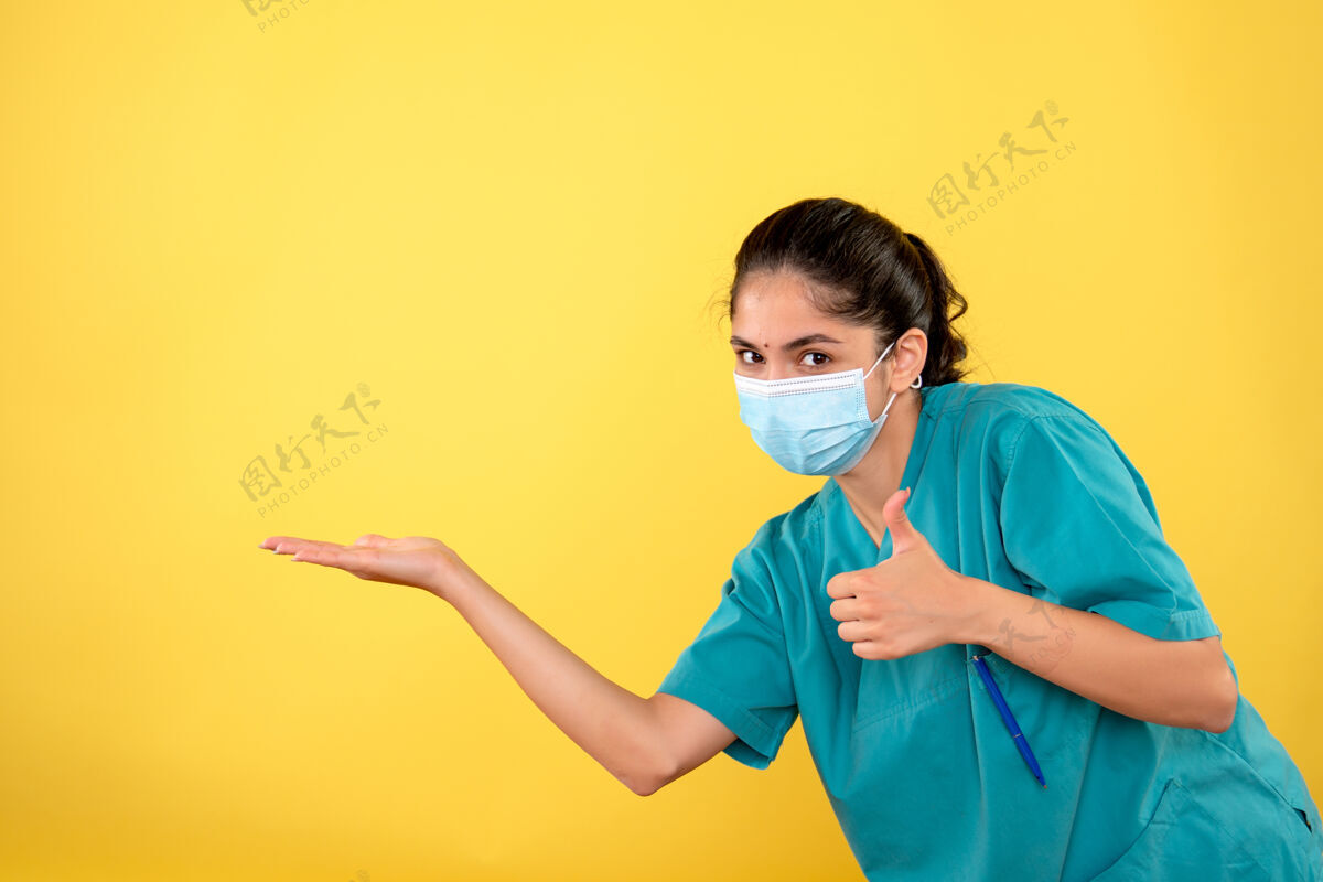 拇指戴着医用面罩的年轻女医生的正面图 黄色墙上有拇指朝上的标志医学微笑成人