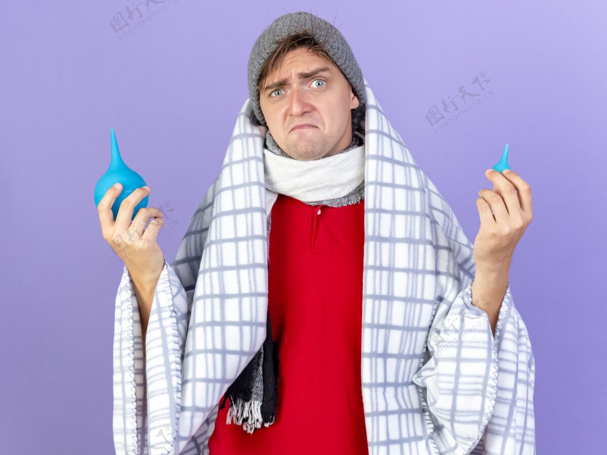 疾病困惑的年轻英俊的金发病男子戴着冬天的帽子和围巾裹着格子布拿着灌肠隔离在紫色的墙上围巾金发格子