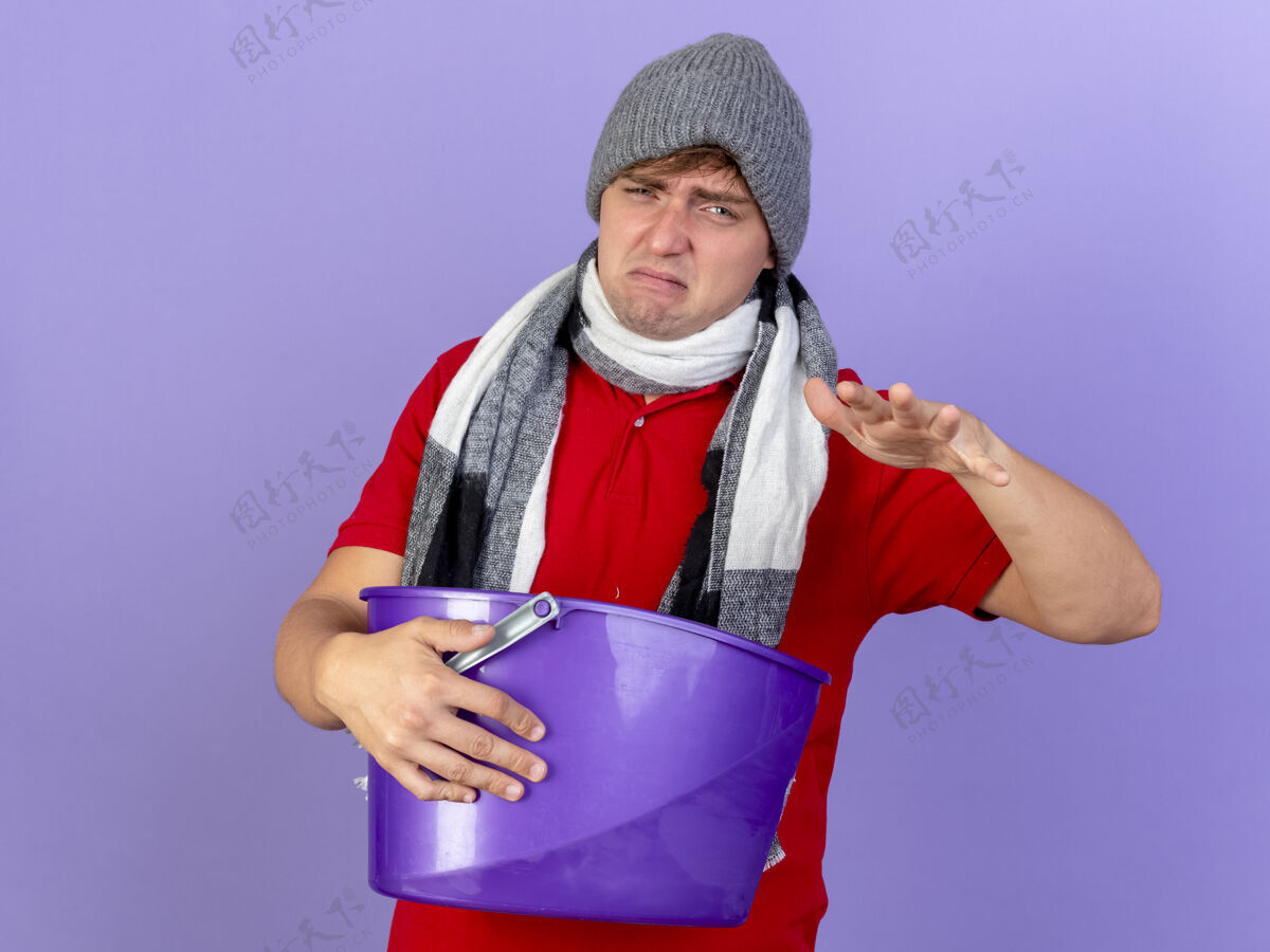年轻年轻英俊的金发病男子戴着冬天的帽子和围巾 手里拿着塑料桶 手伸向空中 隔离在紫色的墙上 留着复制空间疾病表达围巾