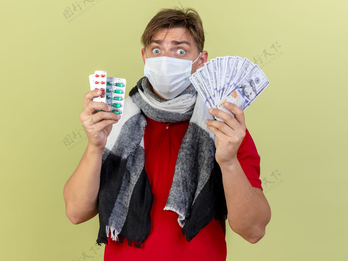 疾病印象深刻的年轻英俊的金发病男子戴着面具拿着钱和医药包隔离在橄榄绿的墙上包装金发年轻