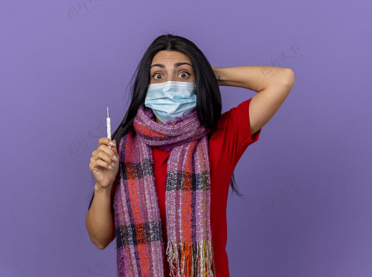 印象印象深刻的年轻白种人生病的女孩戴着面具和围巾拿着温度计 手放在头后 在紫色的墙上与复制空间隔离疾病保持年轻