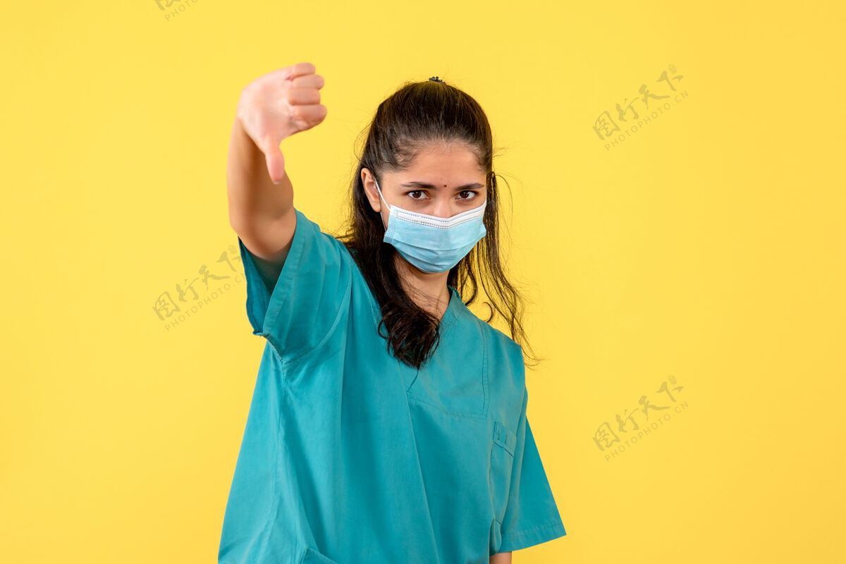 医学黄色墙上戴着医用口罩的漂亮女医生大拇指朝下标志的正面图向下微笑脸