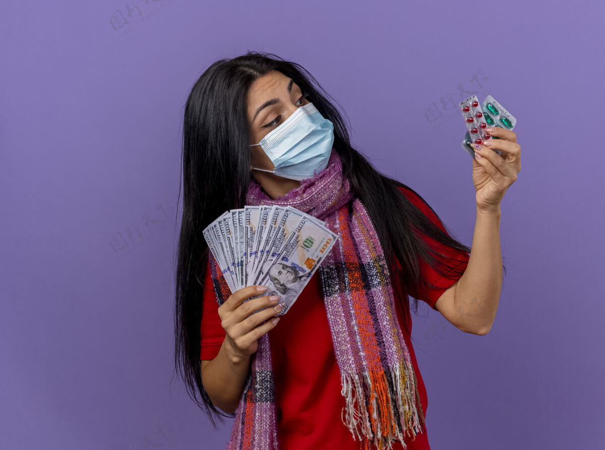 包体贴的年轻白种人生病的女孩戴着面具和围巾拿着钱和一包胶囊看着隔离在紫色墙上的胶囊复制空间疾病穿围巾