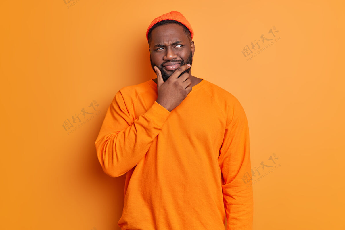 困惑摄影棚拍摄的帅哥留胡子的男人托着下巴若有所思地看着一旁沉思着什么东西戴着帽子和毛衣对着鲜艳的橙色墙壁摆姿势头发解决方案室内