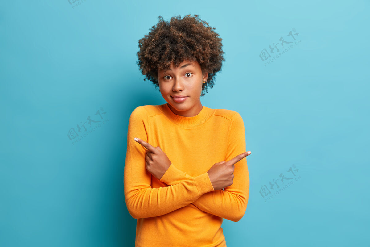美丽可疑的女人交叉双臂 指向不同的侧面 在两件物品之间犹豫不决 或者在蓝色墙壁上摆出橙色套头衫的姿势 需要室内决策台的帮助种族表情手势