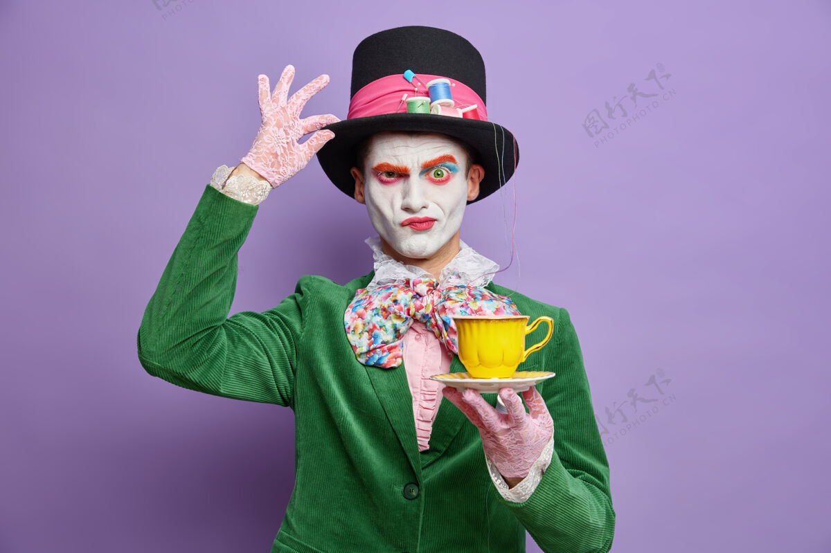 疯狂来自仙境的严肃神秘的男性角色皱着眉头把手放在帽子上喝茶在万圣节的派对礼服上假装疯帽匠有着紫色的隔离墙的彩色化妆品皱眉传统嘉年华