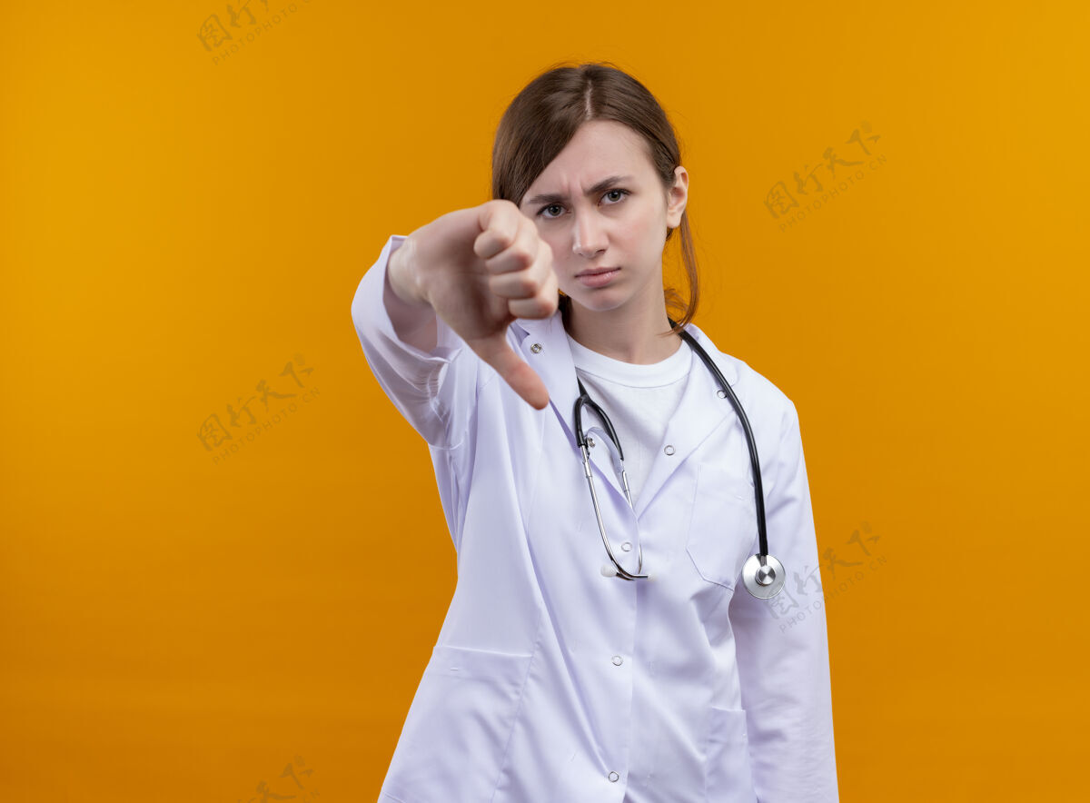显示年轻的女医生穿着医用长袍和听诊器 大拇指朝下放在隔离的橙色墙上 留有复印空间不愉快长袍医生