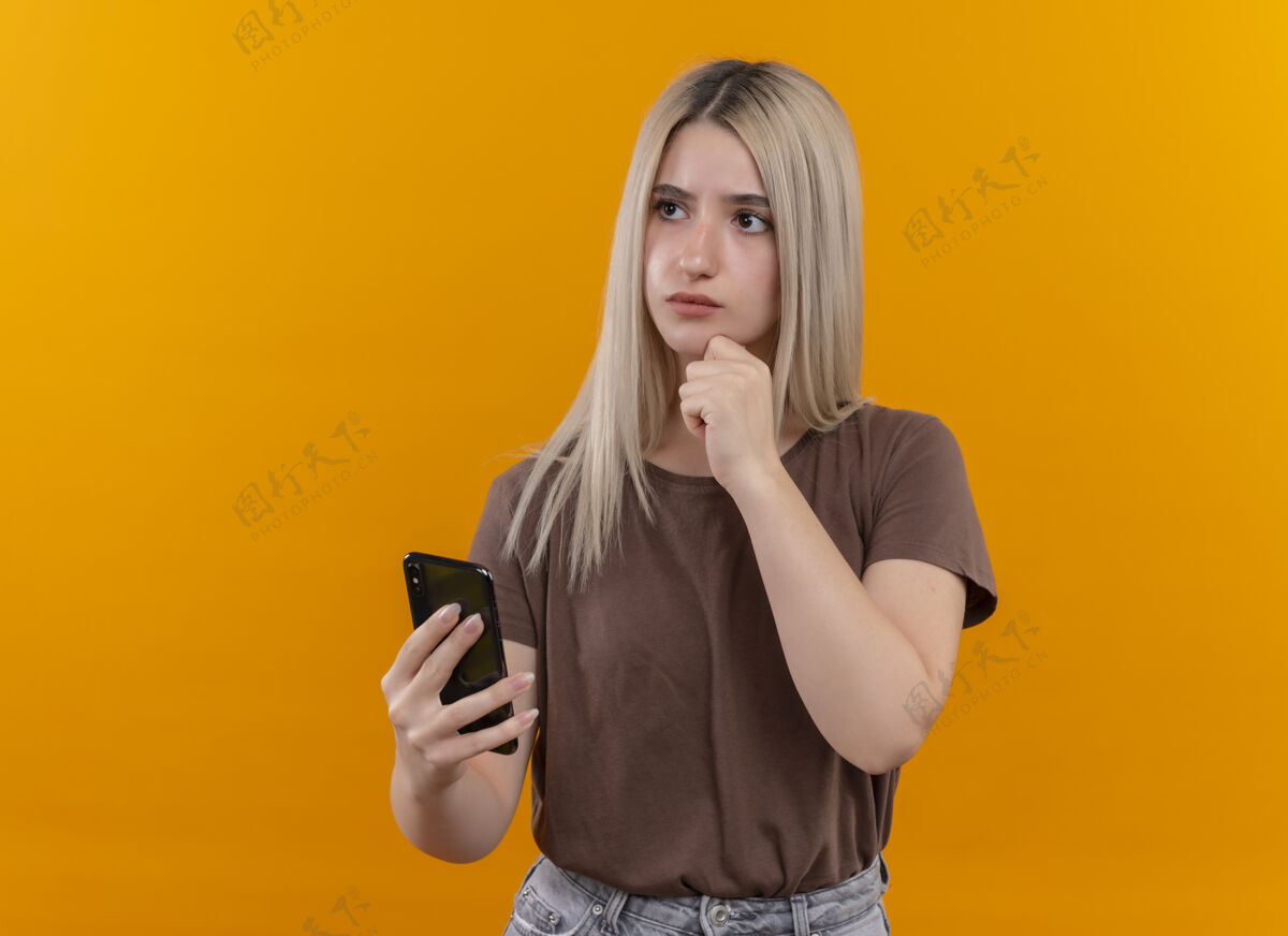 心想体贴的年轻金发女孩拿着手机 手放在下巴上 站在隔离的橙色墙壁上 留着复印空间下巴手机女孩