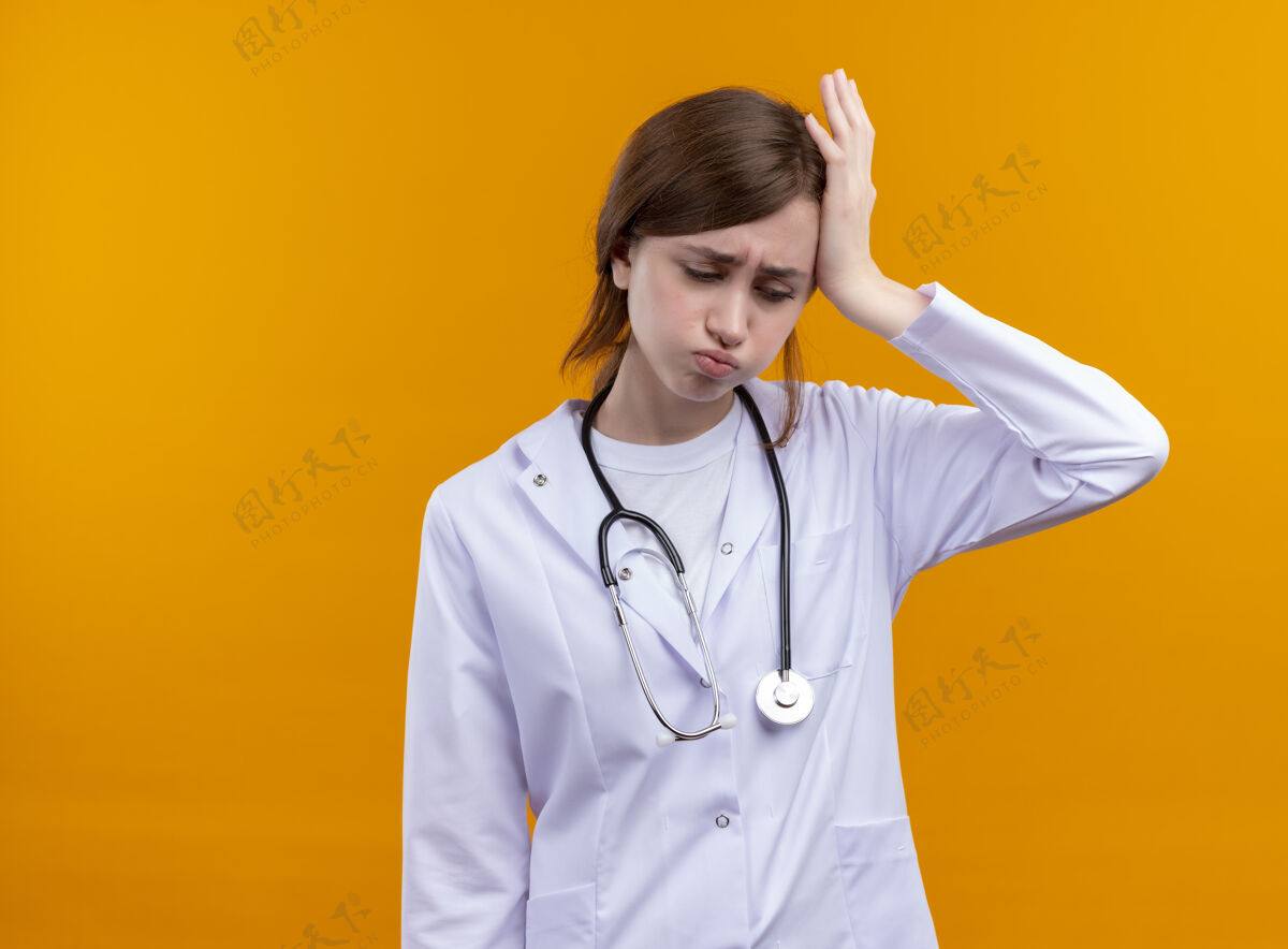 听诊器紧张的年轻女医生穿着医用长袍和听诊器 手放在头上 俯视着隔离的橙色墙壁和复印空间头医生长袍