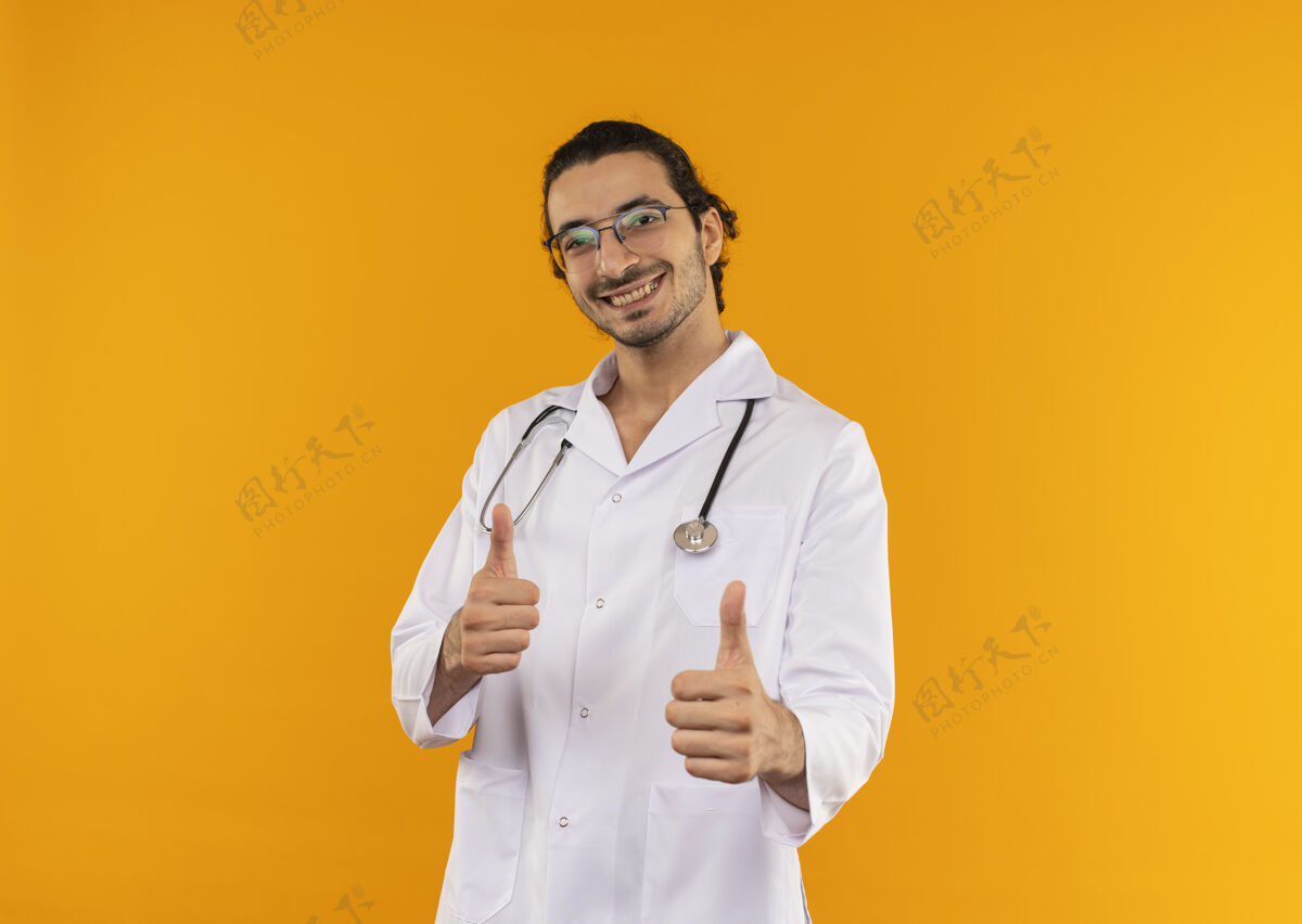 微笑微笑的年轻医生 戴着医用眼镜 穿着医用长袍 带着听诊器 他竖起大拇指站在隔离的黄色墙壁上 留着复印空间拇指医生听诊器