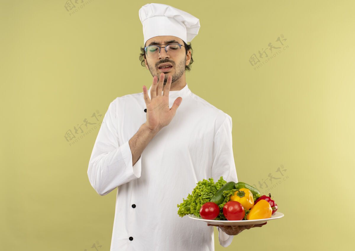 穿闭着眼睛 神经质的年轻男厨师穿着厨师制服 戴着眼镜 把盘子里的蔬菜隔离在绿色的墙上眼镜封闭蔬菜