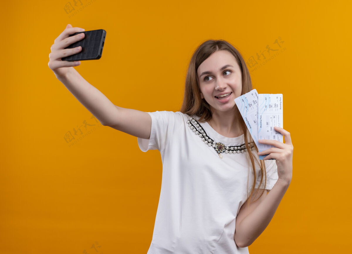 女人微笑的年轻旅人女孩拿着机票 在孤零零的橙色墙上自拍票年轻抱着