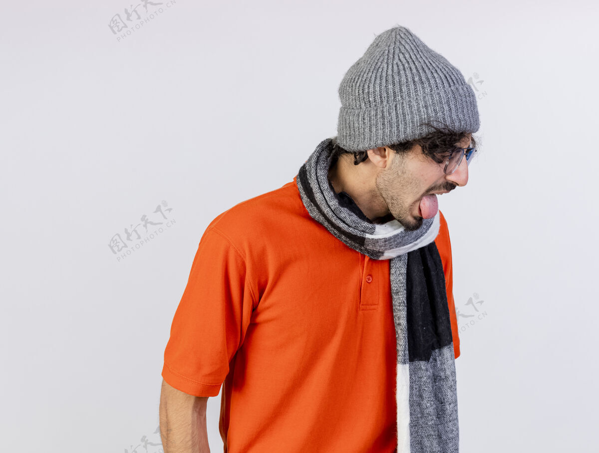 疾病恼怒的年轻白种人 戴着眼镜 戴着冬天的帽子 戴着围巾 把头转向一边 把舌头孤立在白色的墙上 留着复印空间转身舌头烦恼