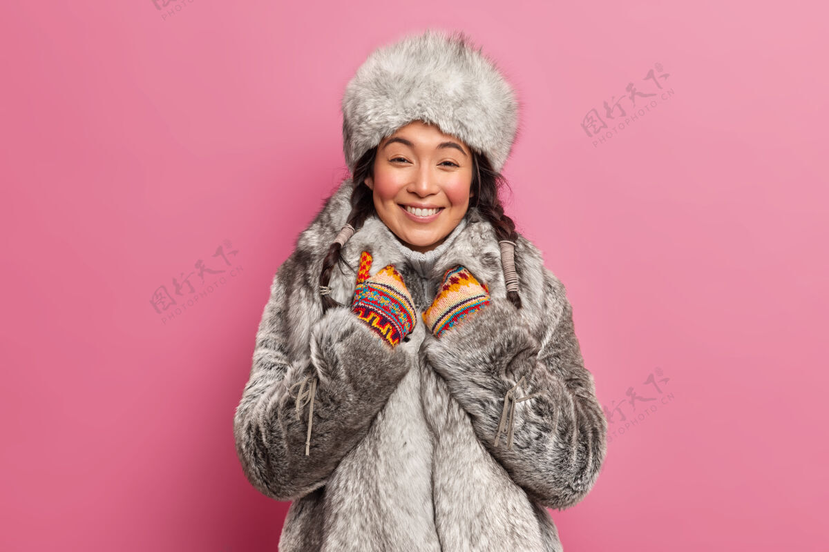 季节穿着灰色皮草外套的漂亮的东方姑娘微笑着真诚地听到有什么愉快的事要在粉色的墙上摆出冬天散步的姿势室内温暖辫子