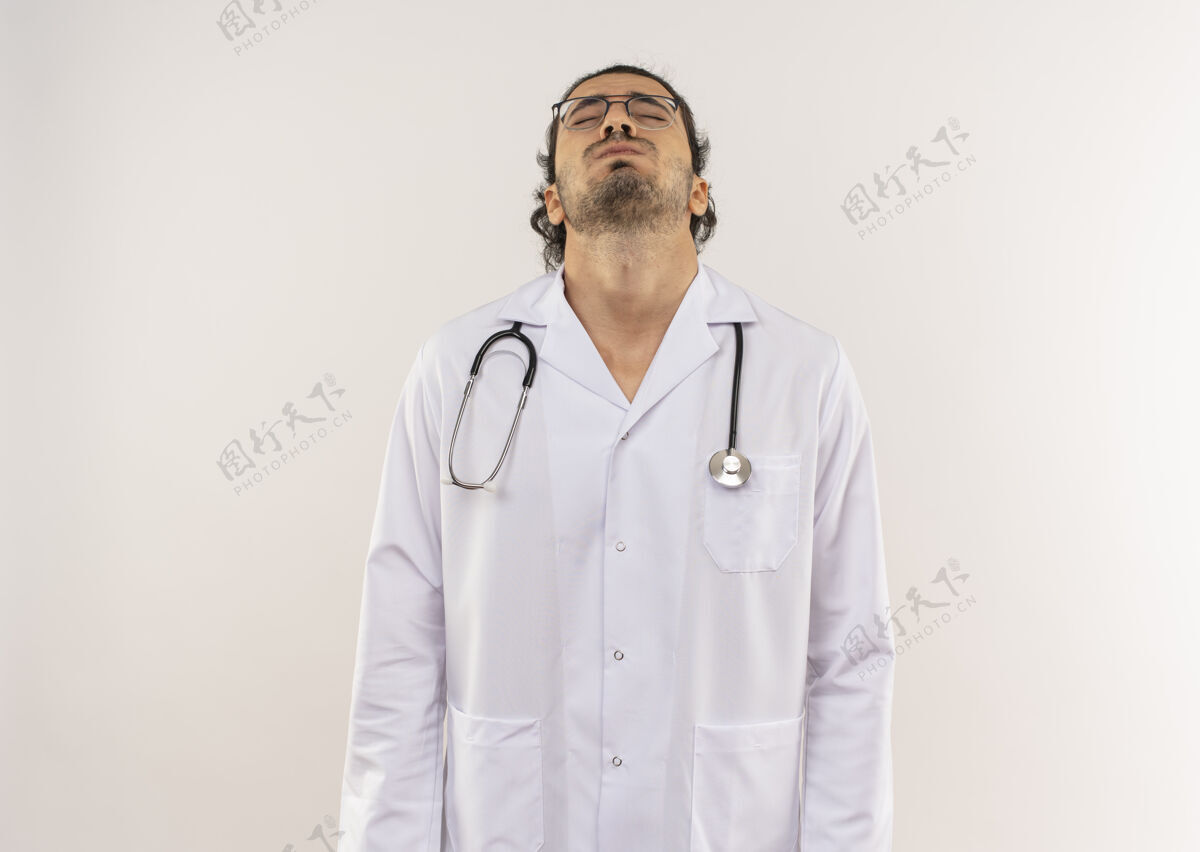 光学闭着眼睛 戴着眼镜的年轻男医生 穿着白色长袍 听诊器 在隔离的白色墙壁上抬起头 留有复印空间提高头医生